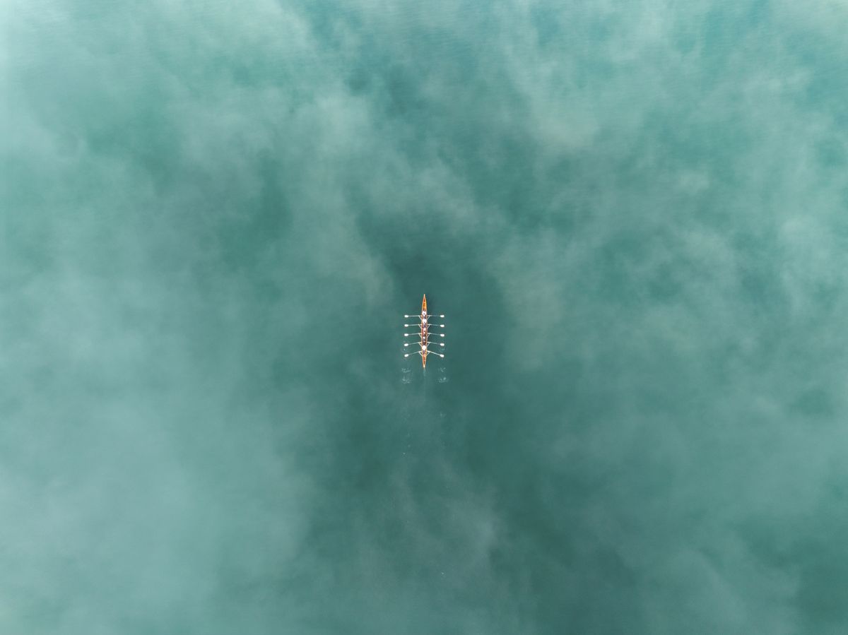 An aerial view of a rowing team gliding through the pristine waters of Lake Zurich in Switzerland. The team is perfectly synchronized, with oars cutting cleanly into the turquoise, misty surface