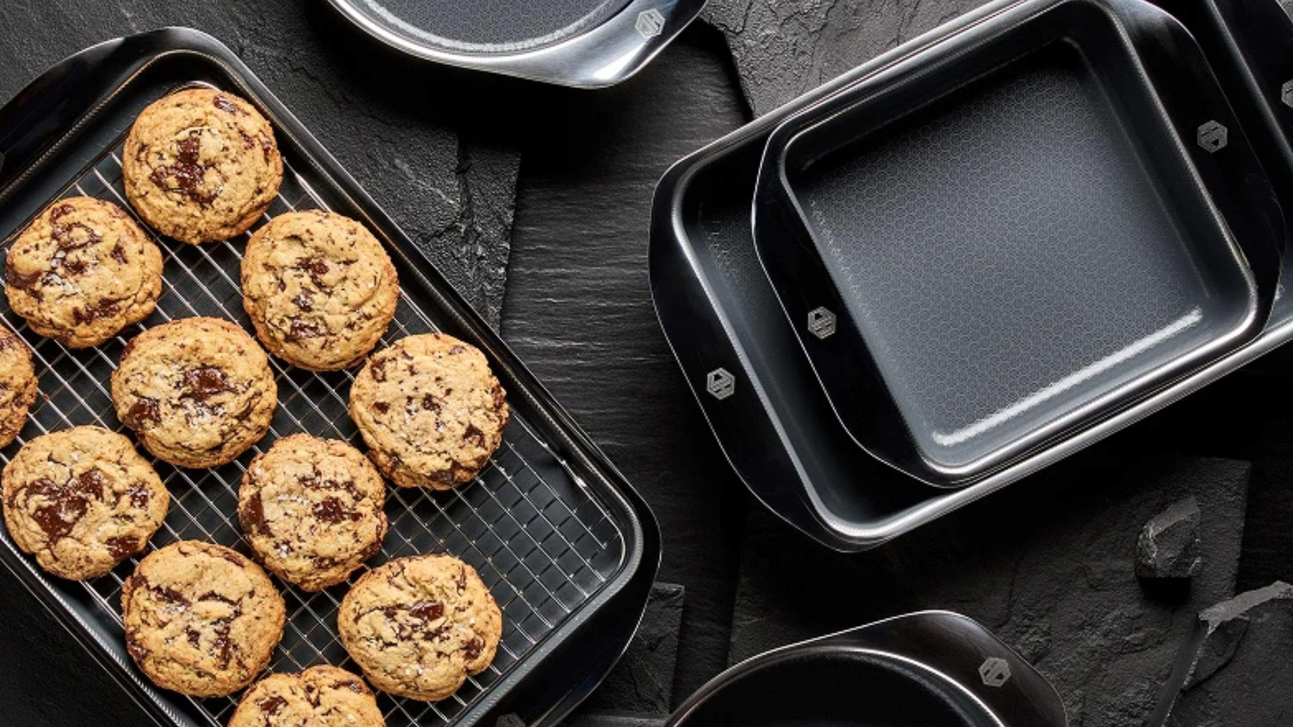 A selection of black HexClad baking trays and tins, one sheet with fresh cookies on top, on a black kitchen counter viewed from above. 