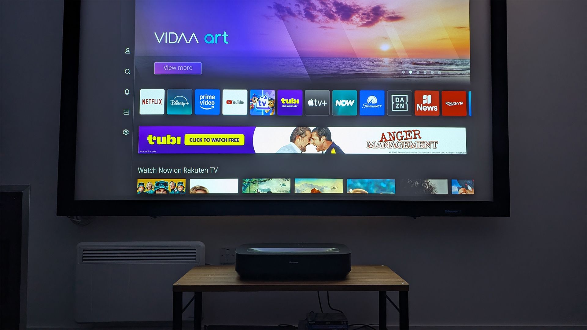 Hisense PL2 UST projector on wooden AV rack in test room. On screen is VIDAA OS home screen