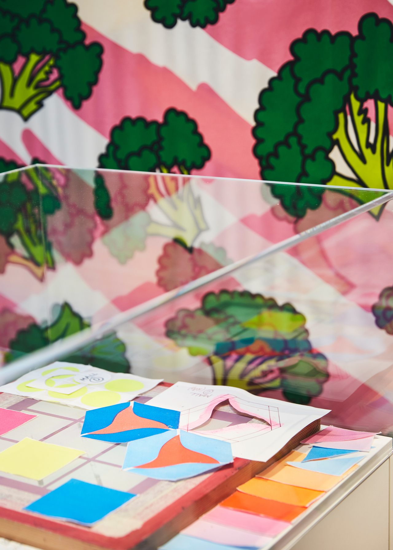 A transparent display case with scraps of color swatches and patterns in front of a fabric with pink and white streaks and green broccoli motifs