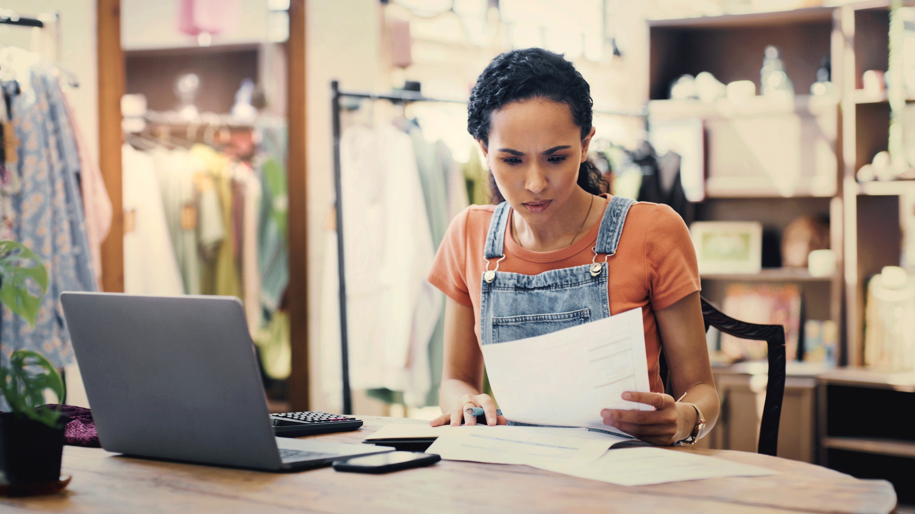 A business owner looks over paperwork at a table in her clothing shop, her laptop open in front of her. 