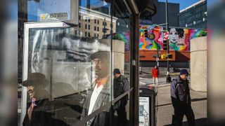 A busy street corner seen through the glass of a bus shelter, its reflections layering a smiling woman in a brown hat, a man in a flat cap and a giant black-and-white advertisement face over a mural