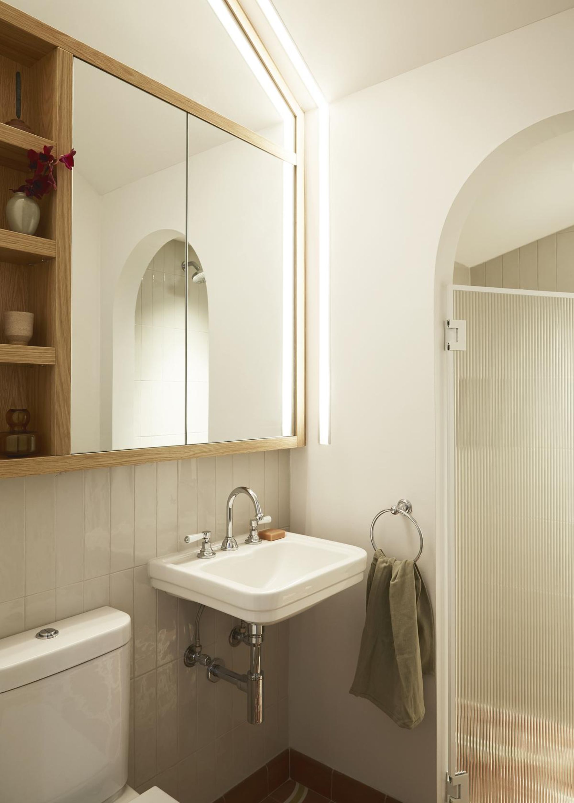 A bathroom with a large mirror cabinet above a small sink. The cabinet also has an open shelving area with essentials inside of it. There is also a zoned area with an archway and glass for the shower room.