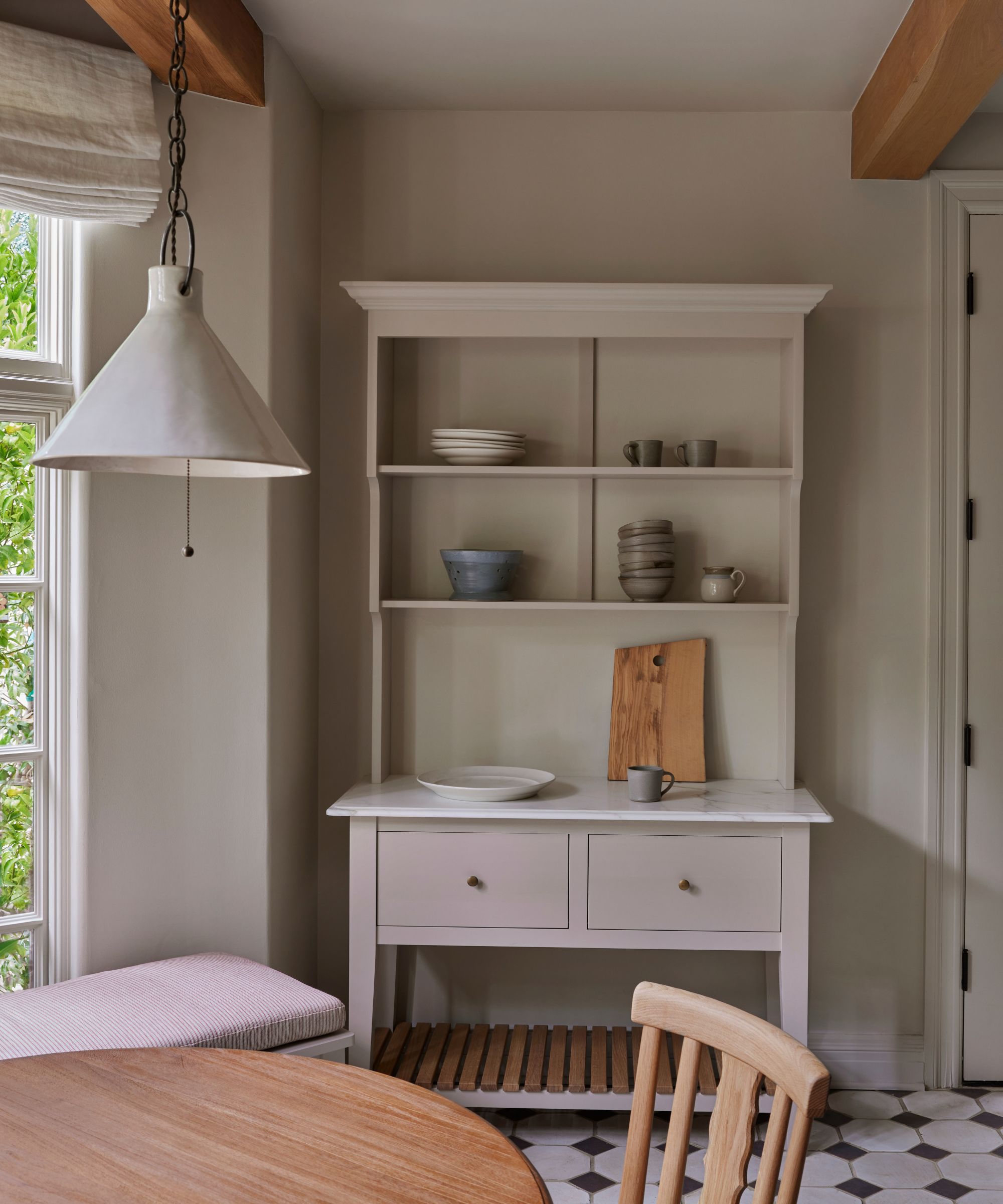 Freestanding painted kitchen hutch with open shelving, drawers and a marble top, styled with ceramics beside a window and dining table.
