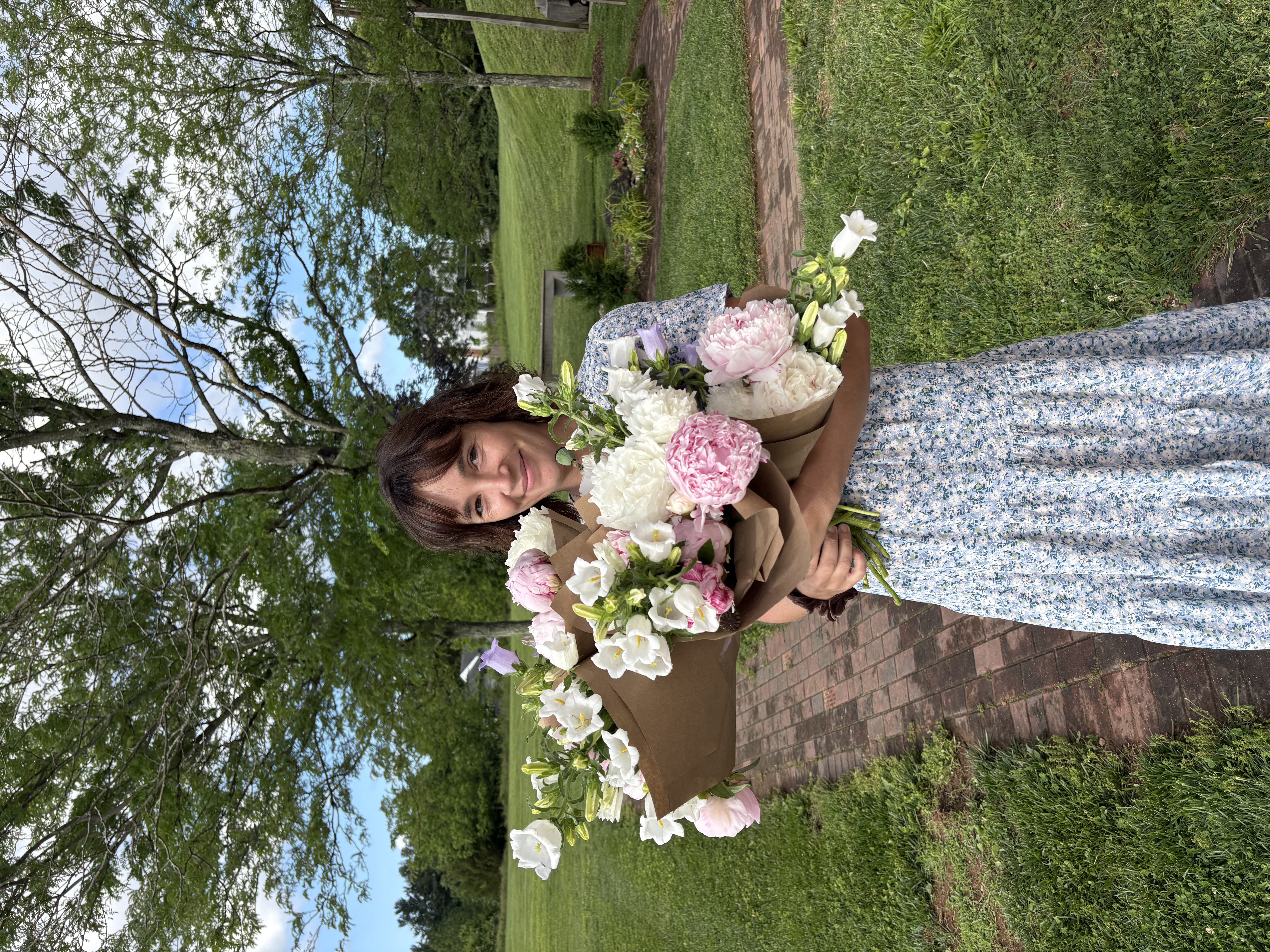 Outdoor shot of Caroline Maurer, head designer at Figment Cottage Gardens wearing blue floral dress and holding large flower arrangements