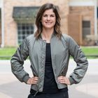 Woman standing outside with her hands on her hips looks at the camera. She is wearing a black T-shirt and a green jacket.
