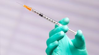 A close-up image of the hand of a person wearing greenish blue lab gloves and holding a vaccine syringe.