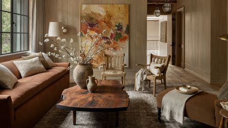 Living room with wooden walls, a rust sofa, a wooden coffee table with rustic vases and branches on top, two wooden woven armchairs, and a brown chaise lounge.