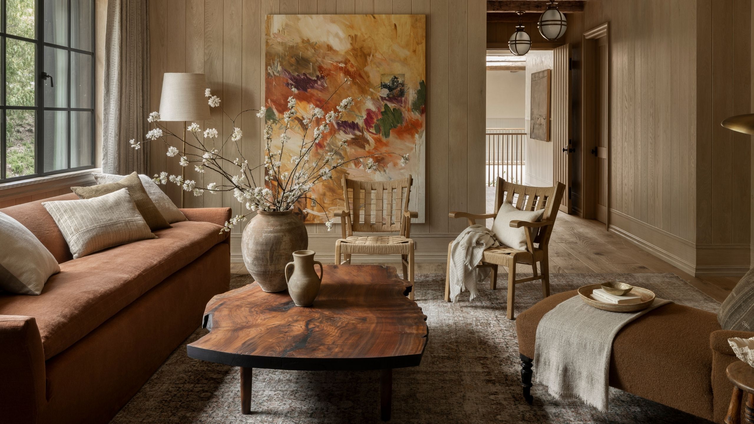 Living room with wooden walls, a rust sofa, a wooden coffee table with rustic vases and branches on top, two wooden woven armchairs, and a brown chaise lounge.