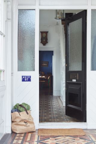 a black front door in a period property, opening up onto the hallway. In the foreground is a tiles porch with a bag of shopping.