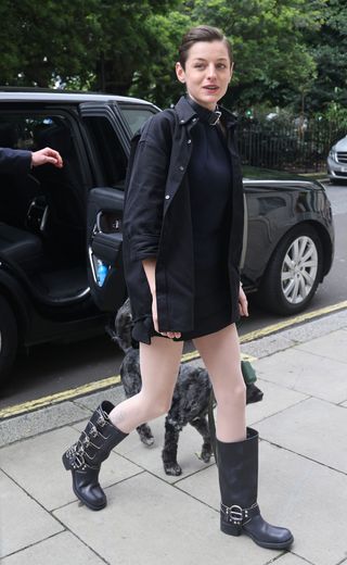 Emma Corrin wears a black mini dress with black buckled boots