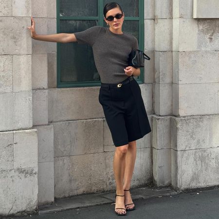 Expensive-looking spring finds for 2025 shown in a photo of a woman standing outside wearing a spring outfit with oval sunglasses, a gray knit top, navy shorts, black strappy sandals, and a black handbag