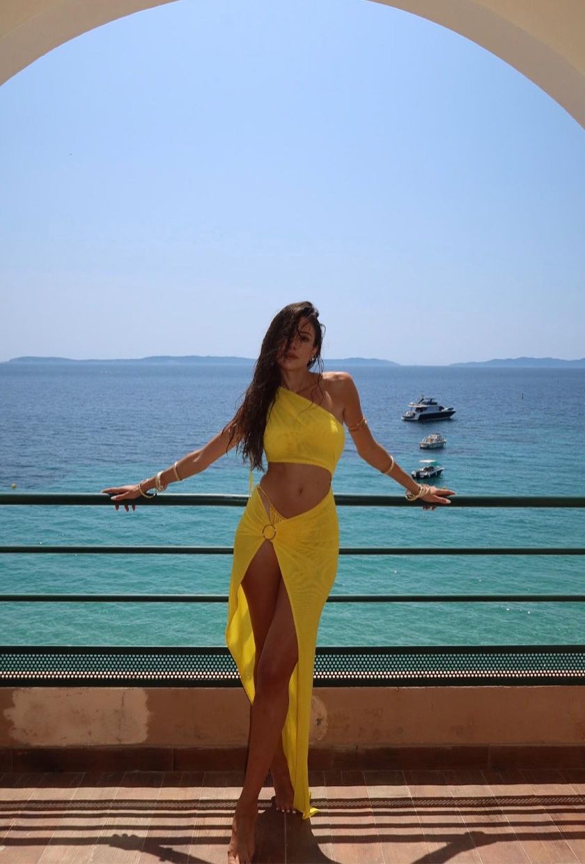 classic swim trend shown on a French woman standing in front of a view of the sea wearing a yellow bikini styled with a matching asymmetrical top and sarong coverup