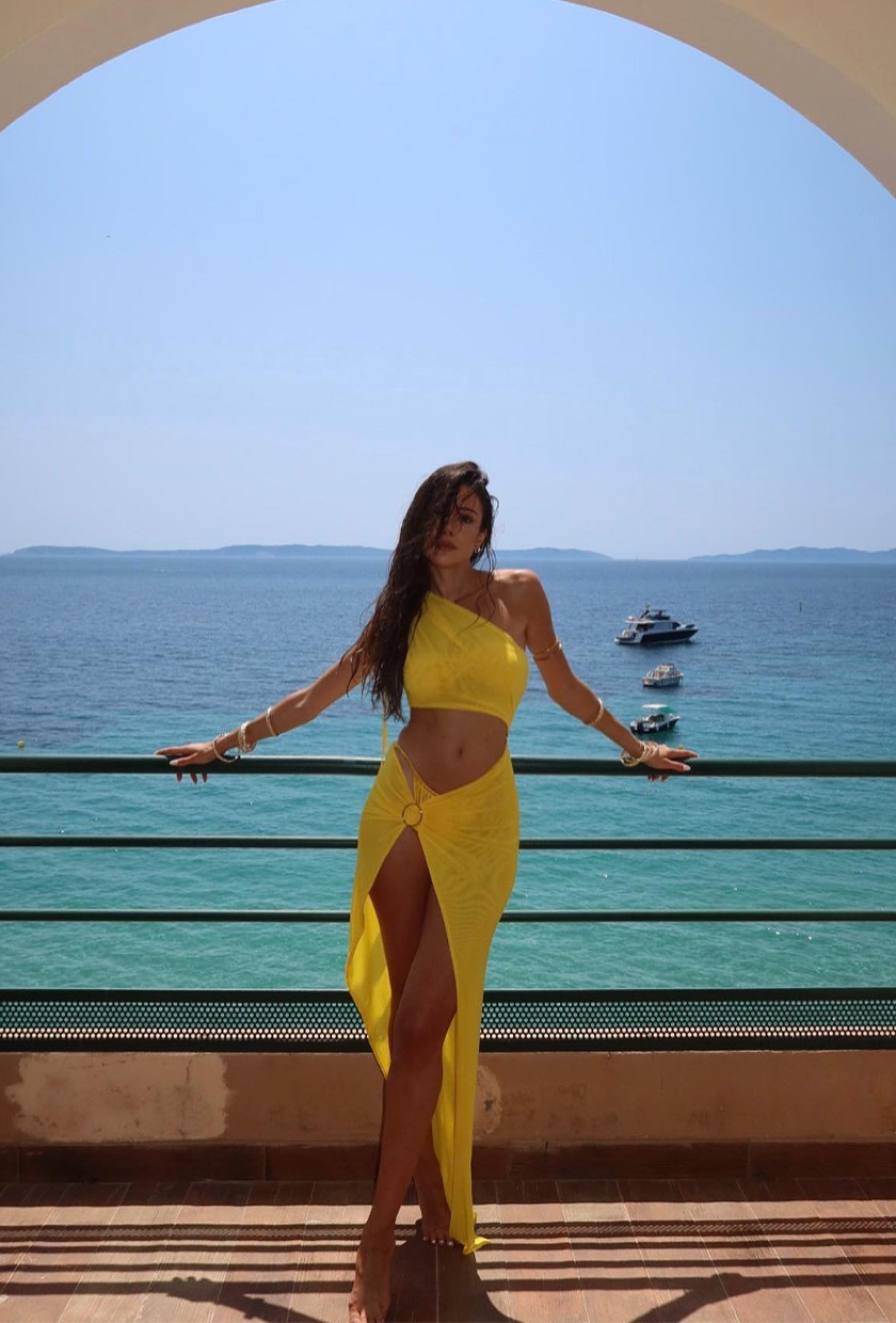 classic swim trend shown on a French woman standing in front of a view of the sea wearing a yellow bikini styled with a matching asymmetrical top and sarong coverup