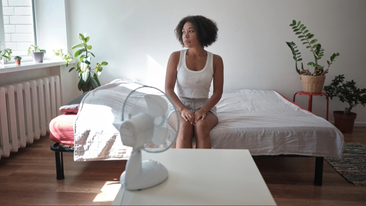 A woman sat on bed with white bedding in bedroom with plants, sunlight coming in through window and a white fan on bedside unit