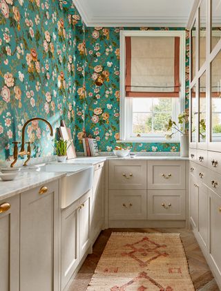 grey galley kitchen with teal wallpaper with blousy pink blooms. White framed window on far wall with white and russet linen blind