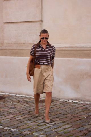 A woman wearing a striped polo shirt, khaki bermuda shorts, pointed toe heels, a brown tote bag, and sunglasses.