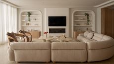 Image of a beige living room painted in an off-white paint color. There is a large, curved, boucle, off-white sectional sofa, two off-white and wood accent chairs, and two beige marble coffee tables. 
