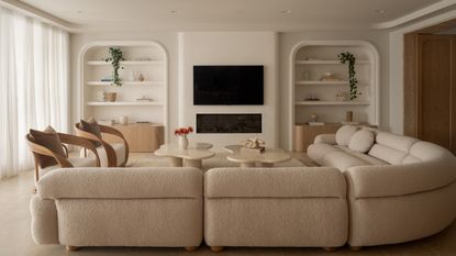 Image of a beige living room painted in an off-white paint color. There is a large, curved, boucle, off-white sectional sofa, two off-white and wood accent chairs, and two beige marble coffee tables.