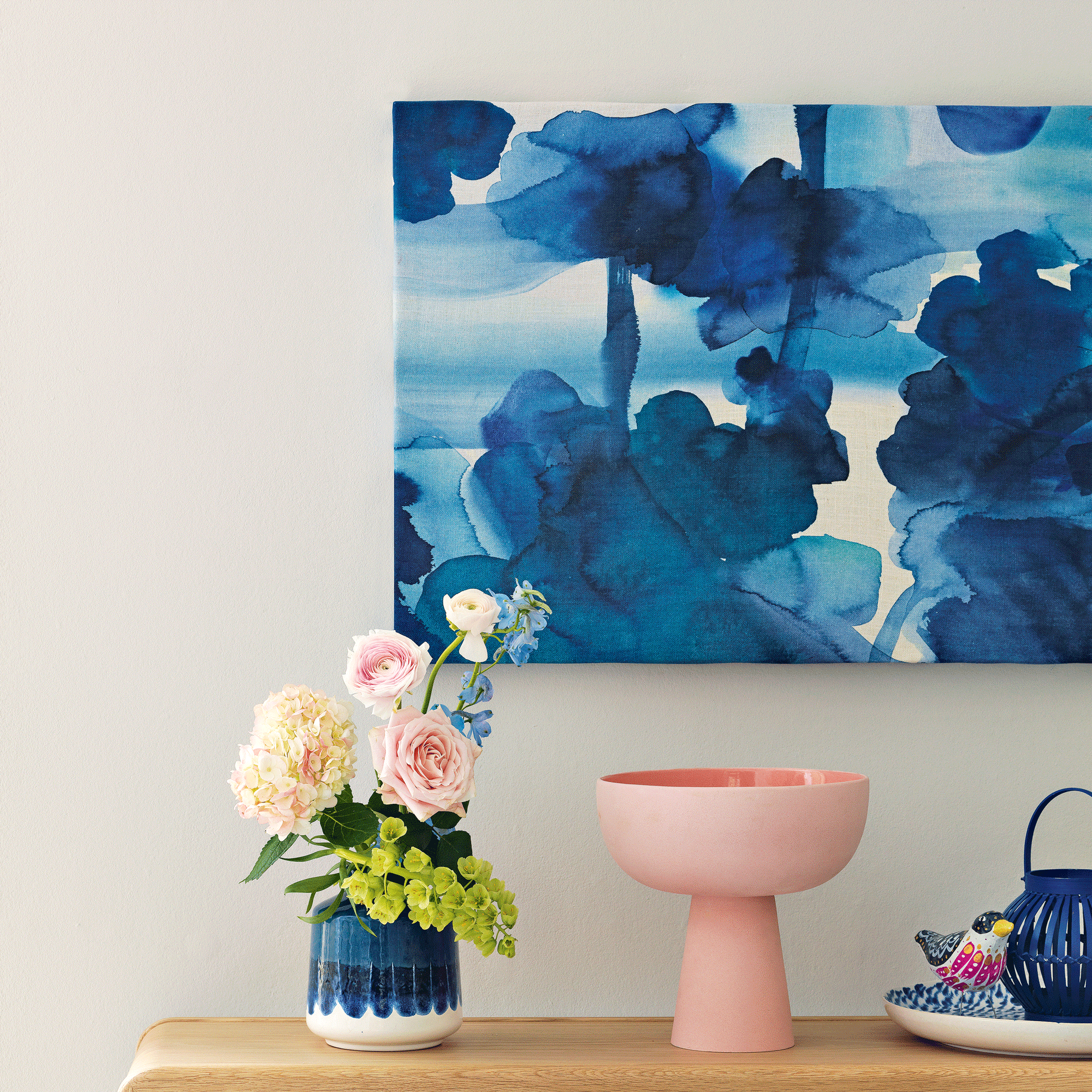 a white wall with as blue and white artwork, with a a sideboard beneath featuring a pink raised bowl vase, a blue and white planter with fresh flowers and a bird decoration