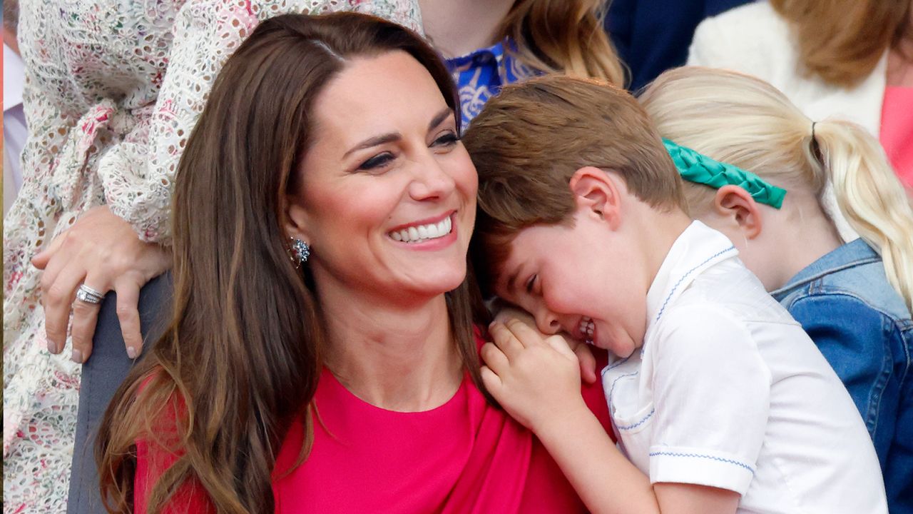 Princess kate wearing a pink dress laughing as Prince Louis puts his head on her shoulder 