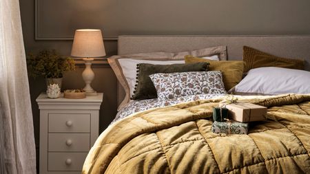 A double bed with a mustard colord velvet throw draped across it. White and green bedding. A white painted side table with four small drawers and a lap on top of it. Some wrapped gifts on the bed. 