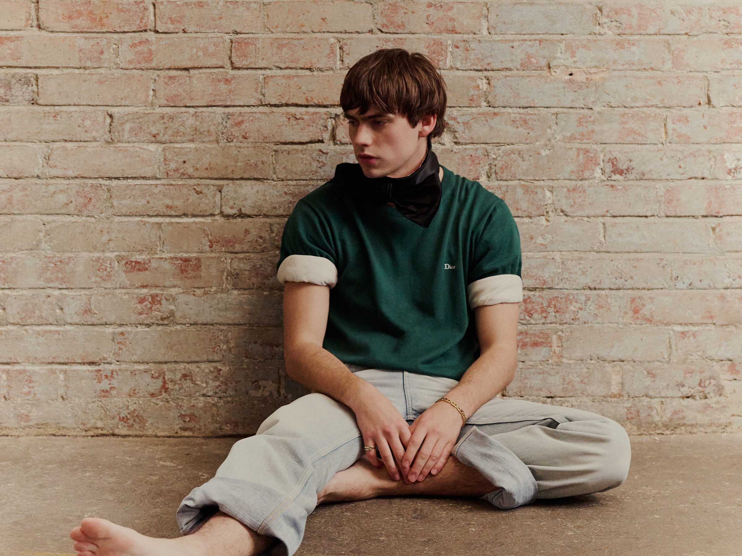 Spike Fearn is sitting on the ground in front of a brick wall wearing a green T-shirt with rolled-up sleeves and cuffed light-wash jeans.