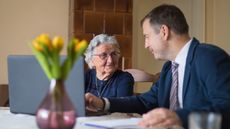 An older woman is helped on her laptop by an elder law attorney at her kitchen table.