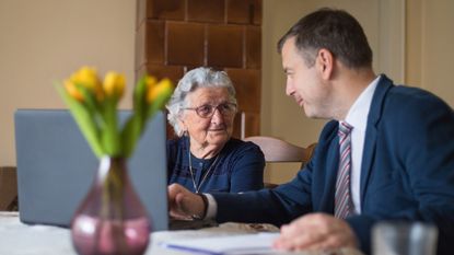 An older woman is helped on her laptop by an elder law attorney at her kitchen table.