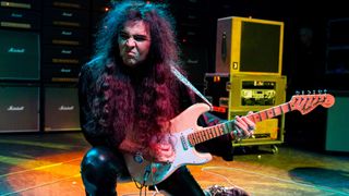 Yngwie Malmsteen kneels onstage as he plays his Fender Stratocaster in front of a wall of Marshall amps