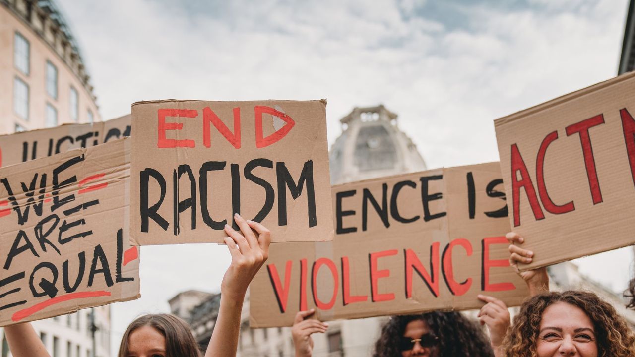 People are marching on strike against racism in the city. They are holding cardboard signs.