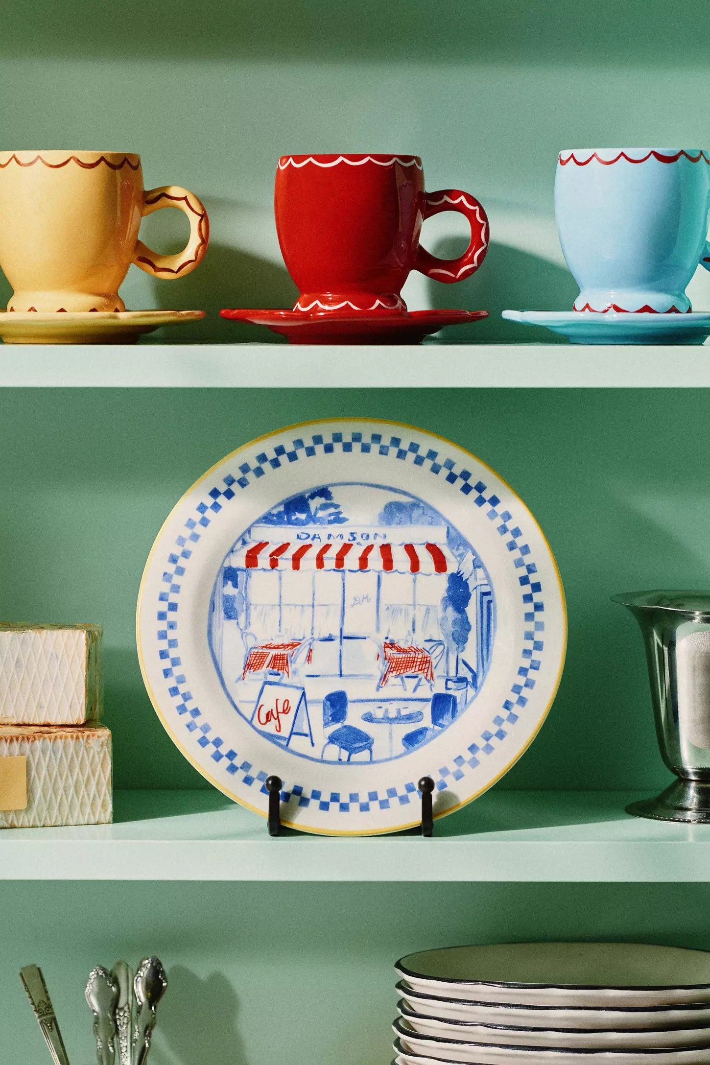 Image of a dessert plate with a cafe motif on it on a mint green shelf. On the shelf above the plate there are three tea cups and saucers in red, yellow, and blue.