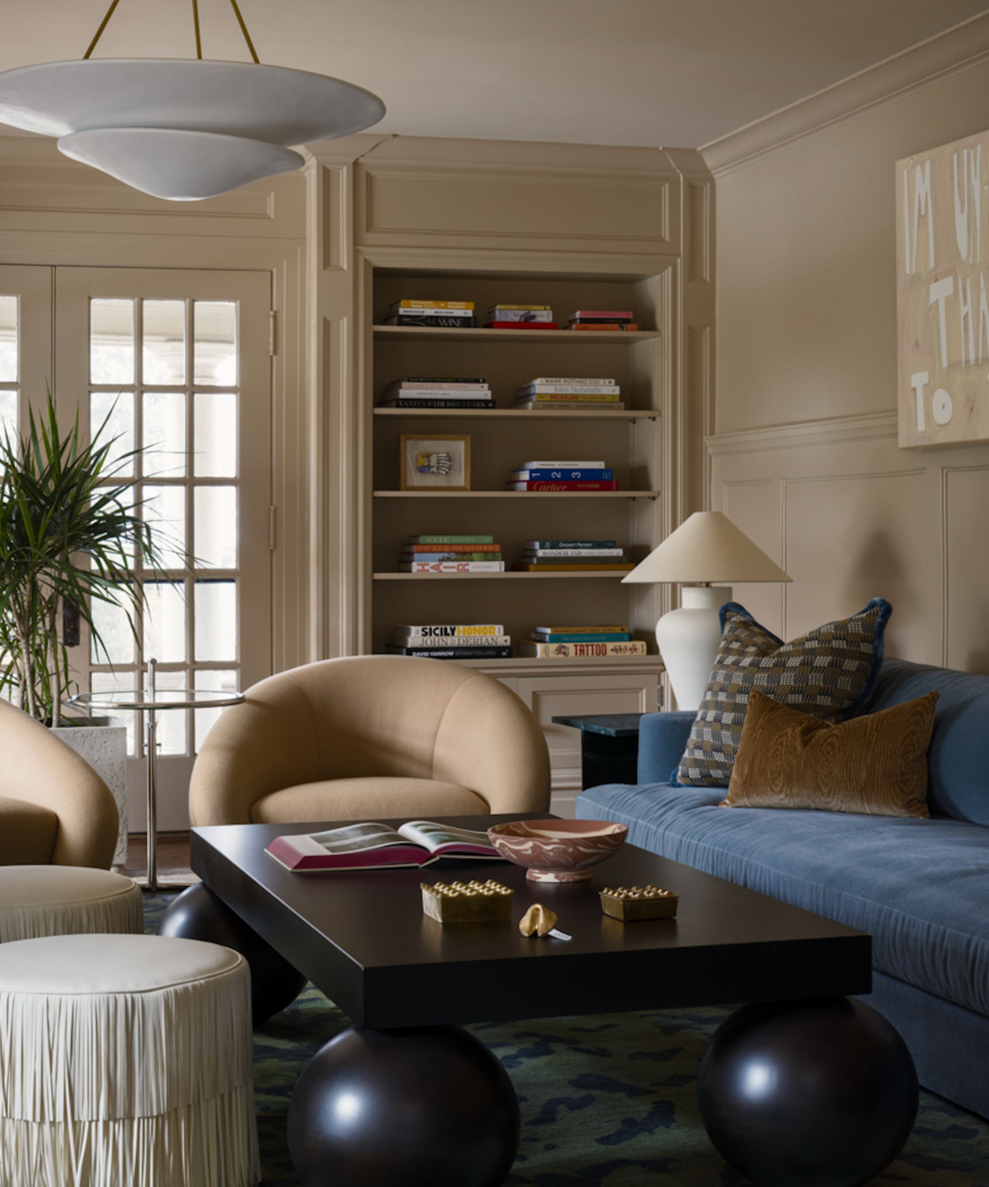 living room with beige walls and built-in shelving, beige armchairs, black sculptural coffee table, and powder blue sofa