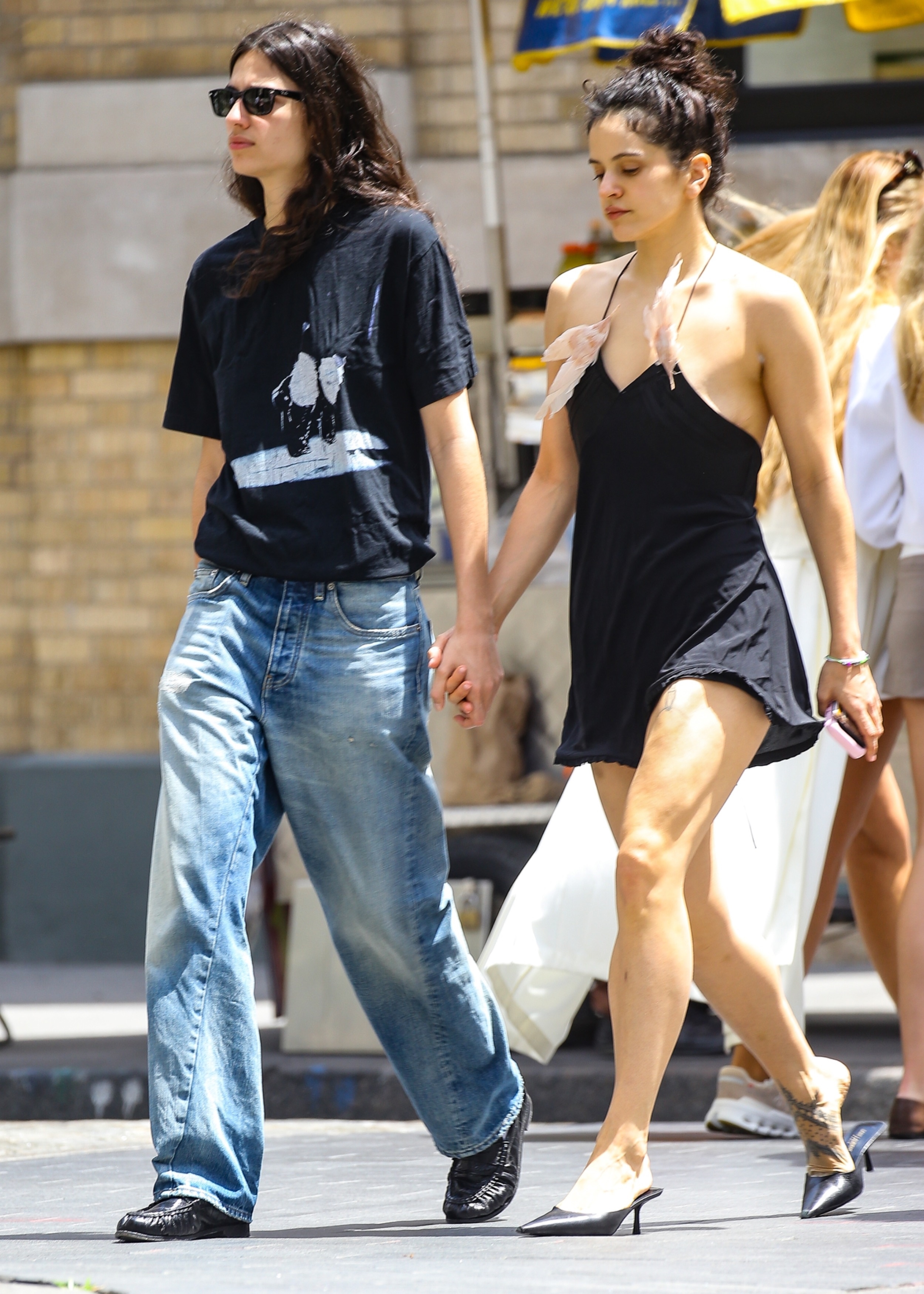 Rosalia wears a black mini dress with two feathers adorned to its straps and pointed-toe mules while holding hands with model Loli Bahia on a date in Manhattan
