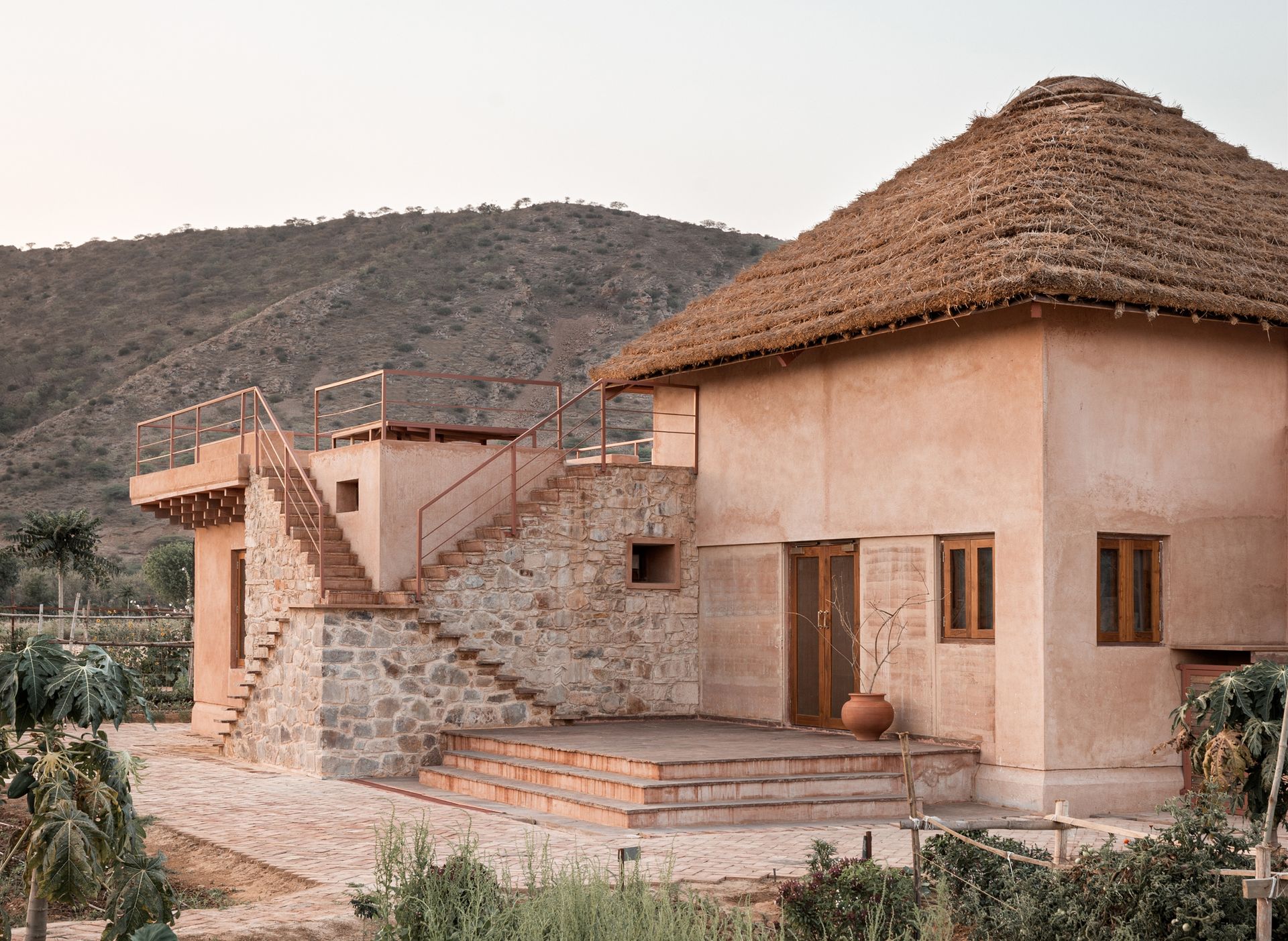 A street level view of a modern-looking beige exterior house with thatched roof and steps leading up to an upper level. Surrounded by hills.