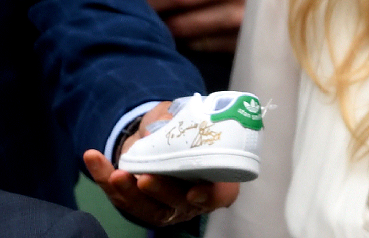 Stan Smith, wearing a navy suit, hands a white Adidas Stan Smith sneaker to Kate Middleton, which he has signed for Prince Louis in gold ink