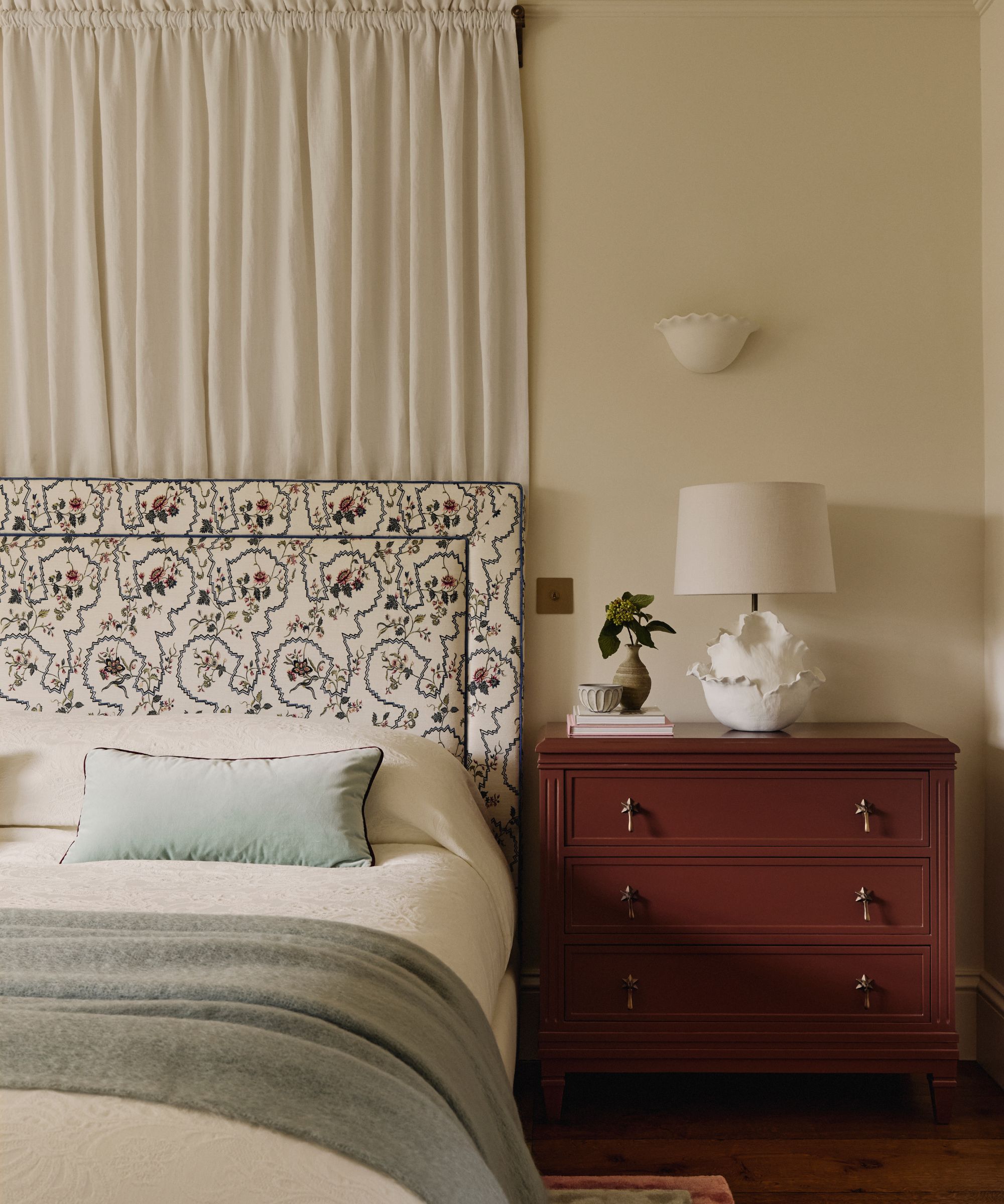 Traditional bedroom with canopy bed, upholstered floral headboard, a mahogany chest of drawers with a white ceramic table lamp on top, and a vase