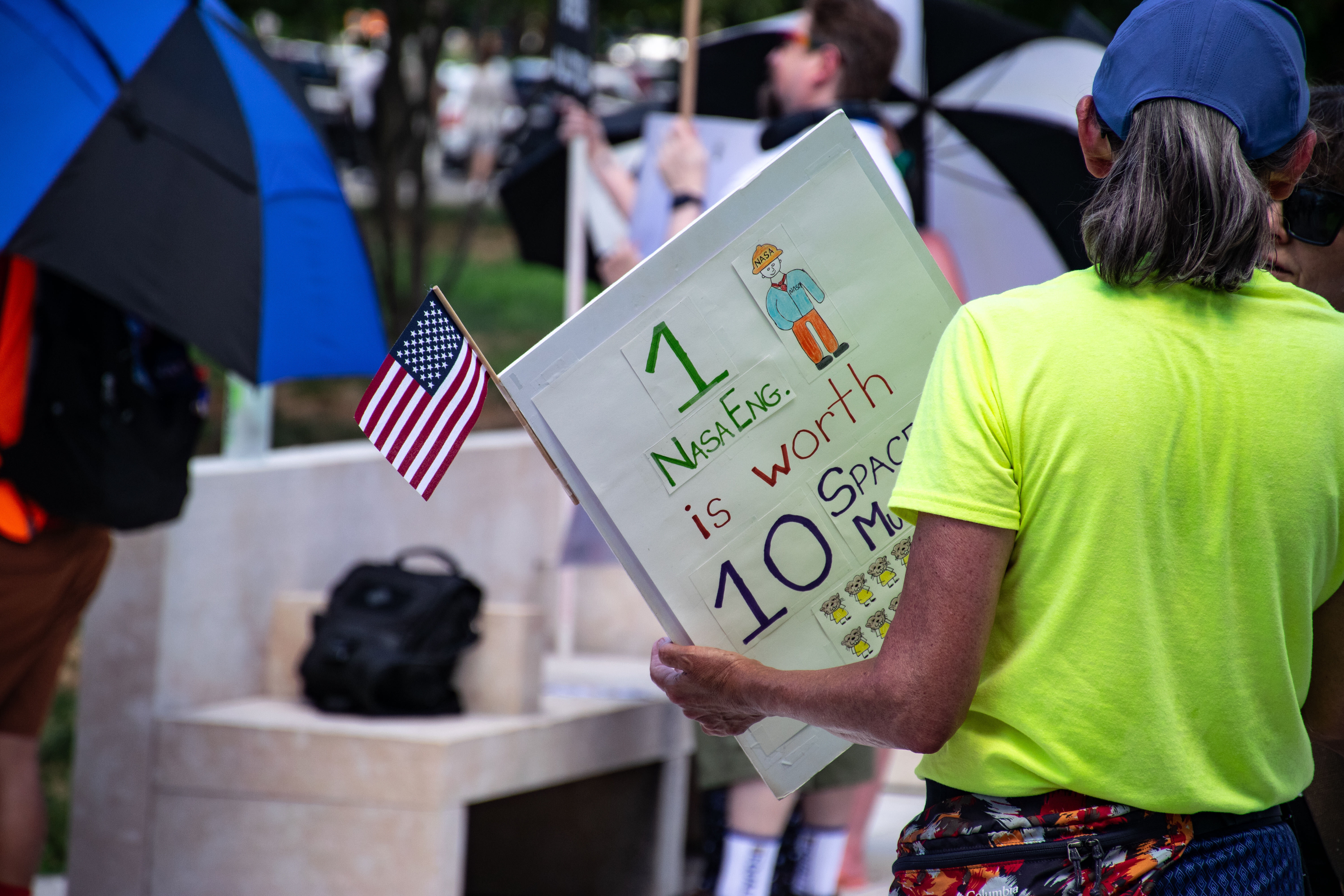 People hold signs outside on a sunny day in protest of NASA budget cuts, July 20, 2025.