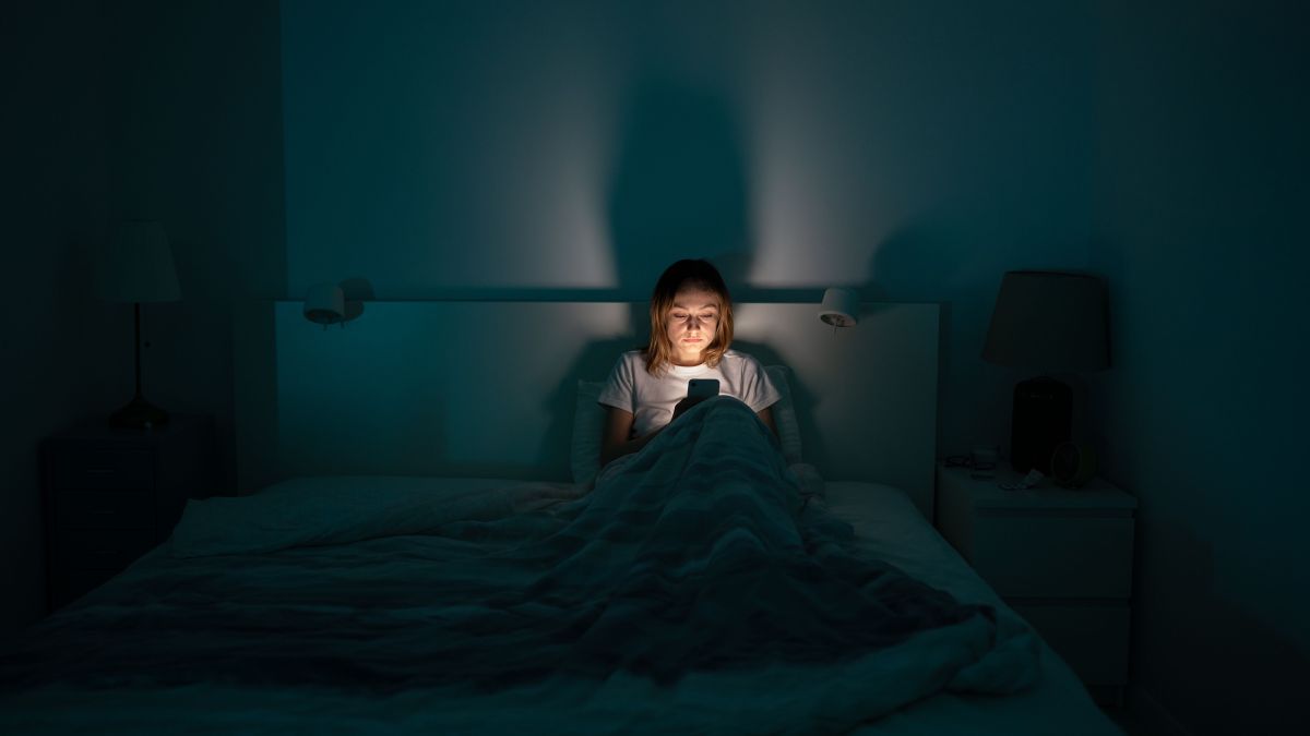 Sleepy exhausted woman lying in bed using smartphone, can not sleep. Insomnia, addiction concept. Sad girl bored in bed scrolling through social networks on mobile phone late at night in dark bedroom.