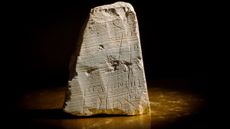We see a triangular gray stone slab sitting that is inscribed with Hebrew against a black background.
