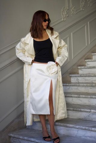 A woman standing on steps wearing a white satin coat over a black tank top with a white satin skirt and black heeled mules.