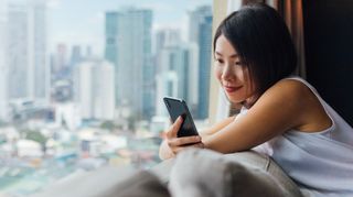 Young Asian woman using smartphone, sitting on the sofa, near the window with city views.