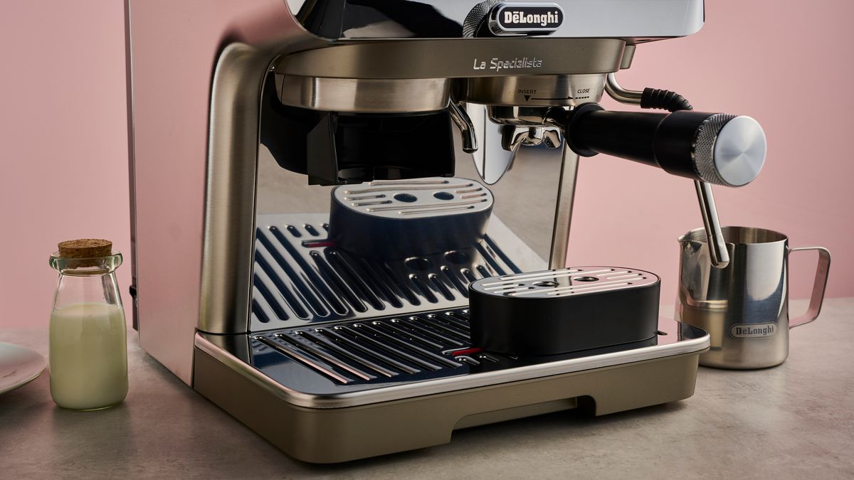 The Delonghi Specialista Arte Evo sits on a stone-effect surface in front of a pink backdrop. It has a small cork-topped bottle of milk to its left, and a metal milk jug to its right.