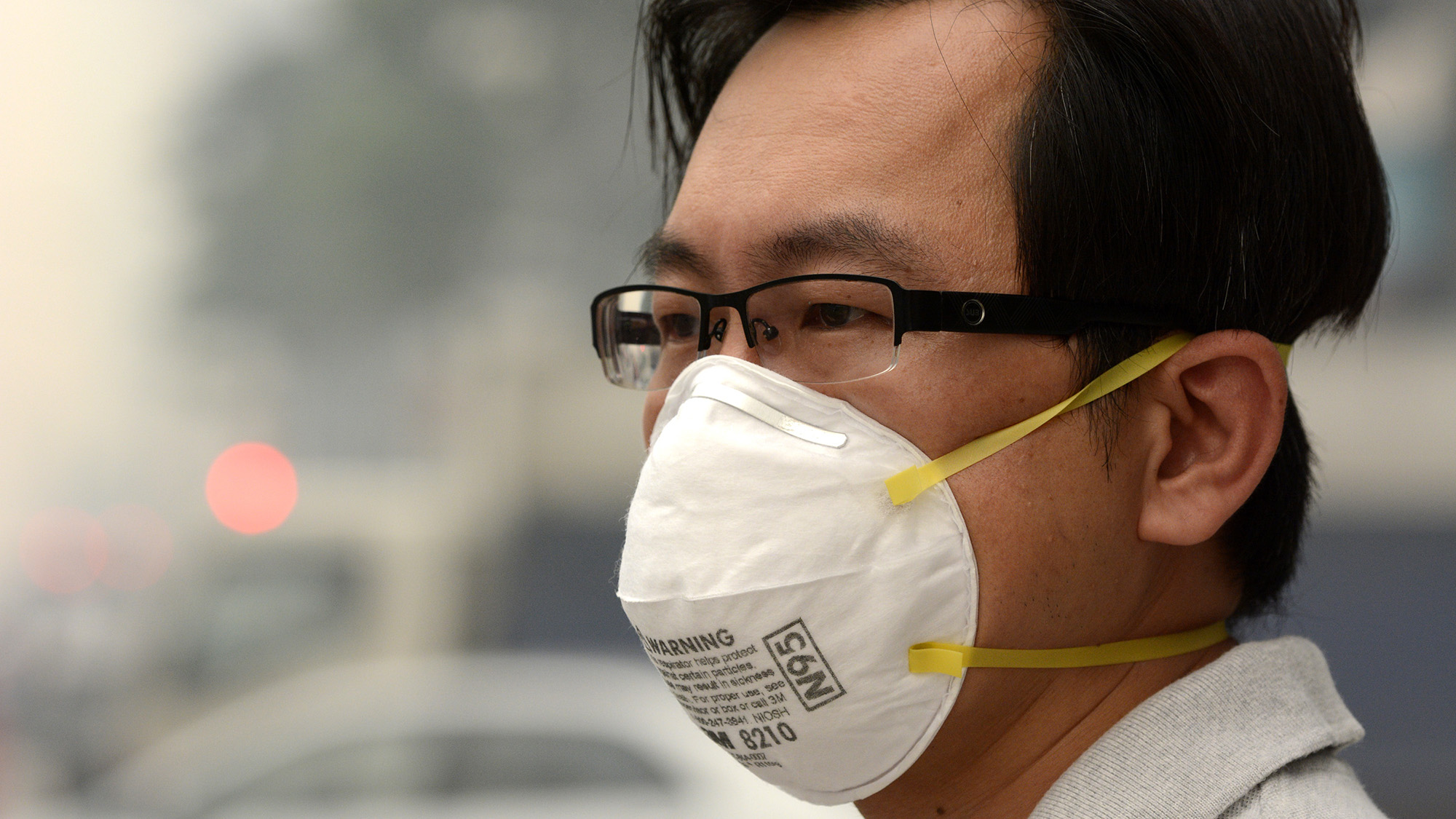 A man wears glasses and a white face mask. Smog is visible in the background.