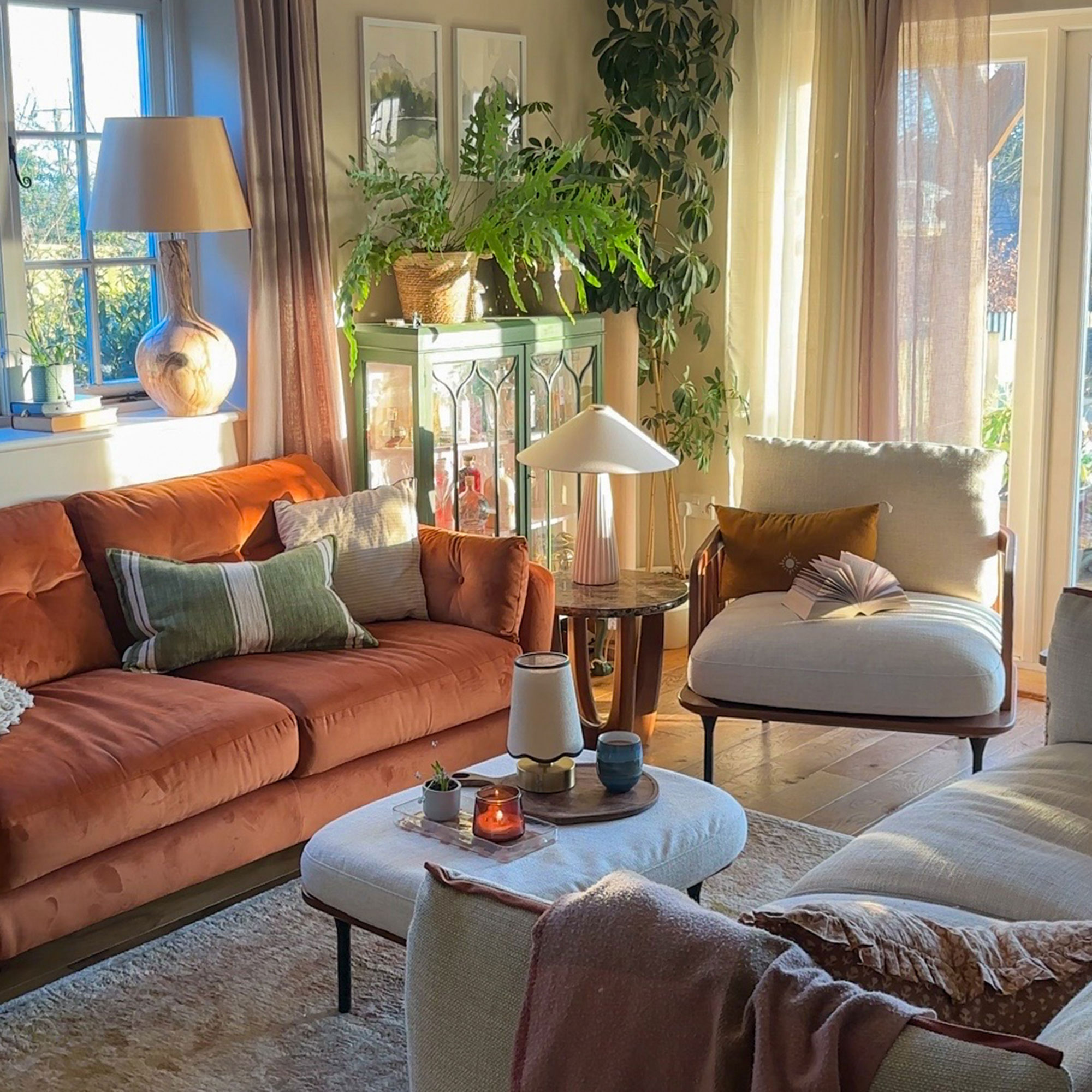 Sunlit living room with burnt orange sofa, cream ottoman as coffee table with matching armchair, green trimmed glass-fronted cabinet, houseplants and sheer curtains