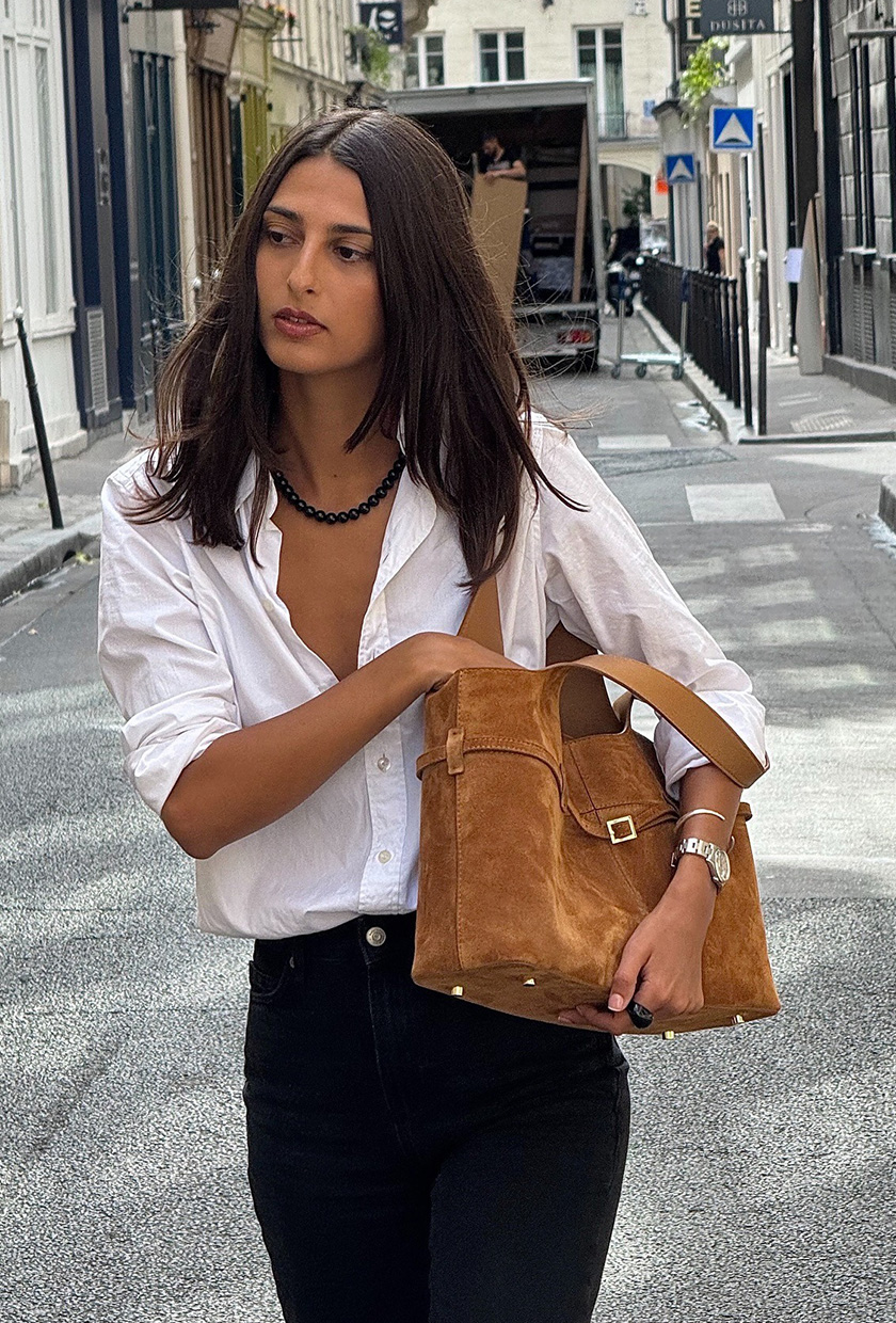 A photo of a woman wearing a white button-down shirt with a green bead necklace, dark wash jeans, a tan suede tote bag, and a silver watch