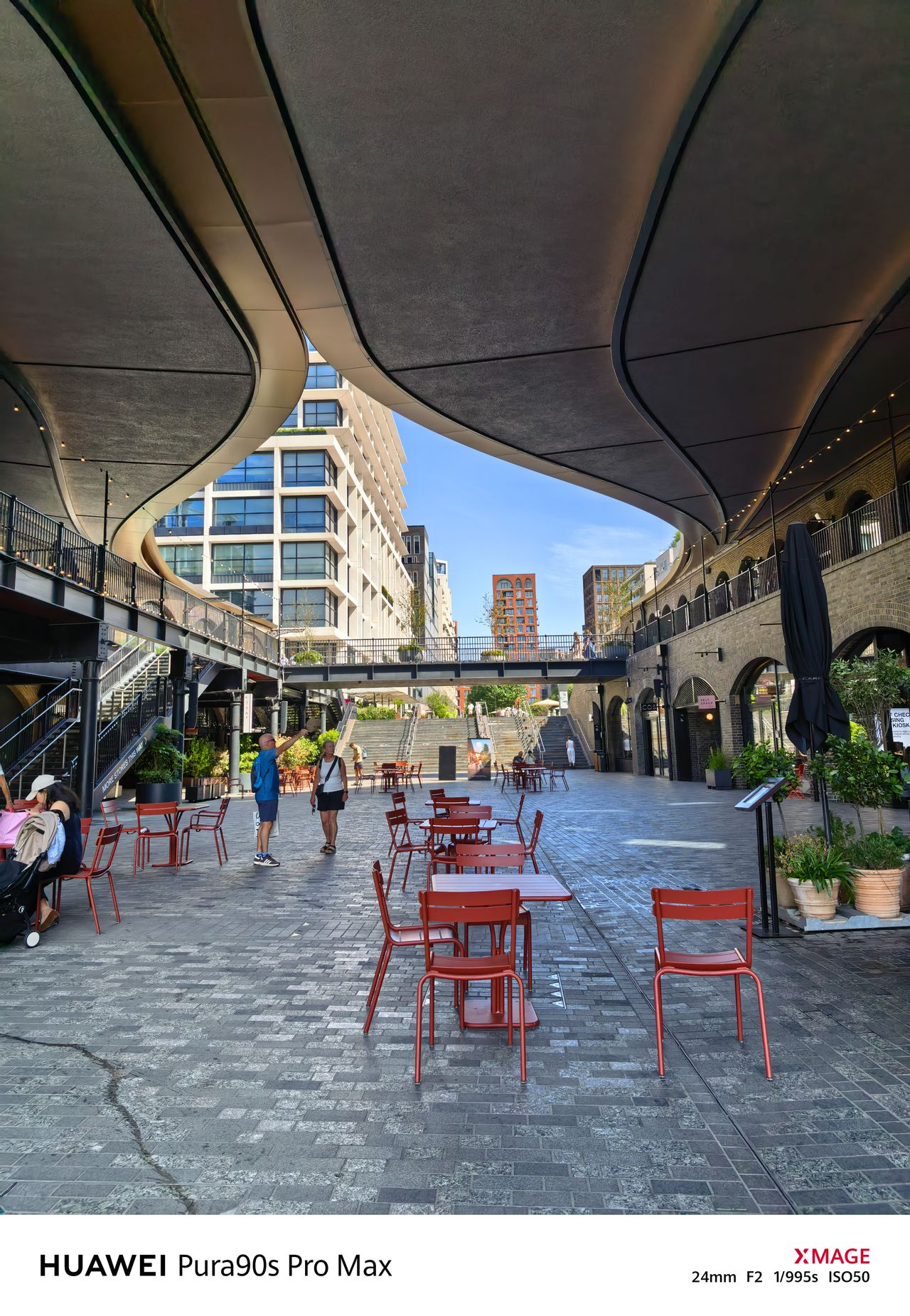 Modern pedestrian courtyard beneath a sweeping curved canopy, with red outdoor tables, chairs and a few people.