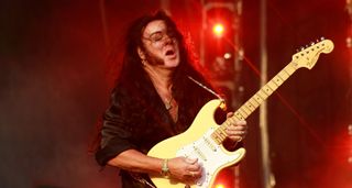 Yngwie Malmsteen takes a solo on his Fender Strat, on a stage bathed in blood-red stage lights.