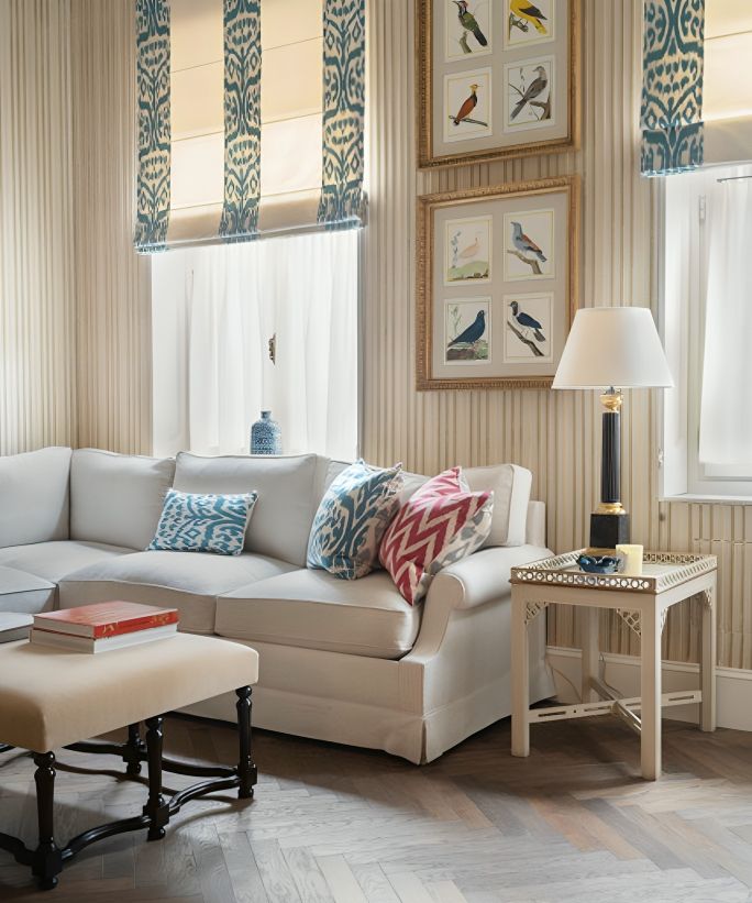 A living room with a brown wood Herringbone floor, a cream sofa, and yellow and white stripped wallpaper. Blue and white roman blinds on large windows, and two large square framed prints on the wall above the sofa.