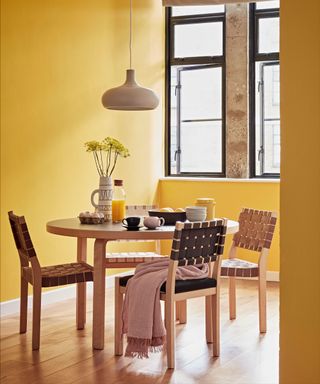 Dining room painted a bright yellow, wooden rounded dining table with four wooden chairs with webbed seat and back, wooden flooring, white low hanging pendant over dining table, table decorated with kitchenware and vase